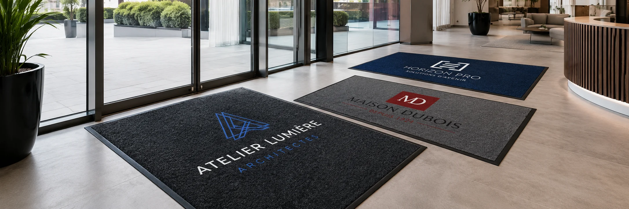Banni&egrave;re r&eacute;aliste pr&eacute;sentant plusieurs tapis d&rsquo;entr&eacute;e professionnels personnalis&eacute;s avec logos fran&ccedil;ais dans un hall d&rsquo;accueil moderne, pour illustrer les solutions de tapis logo sur mesure propos&eacute;es par Kom&eacute;r&egrave;s aux entreprises.